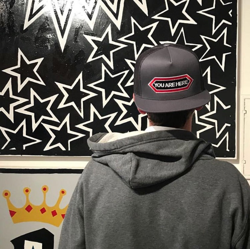 Person wearing a cap with 'YOU ARE HERE' text in front of a wall with star patterns.
